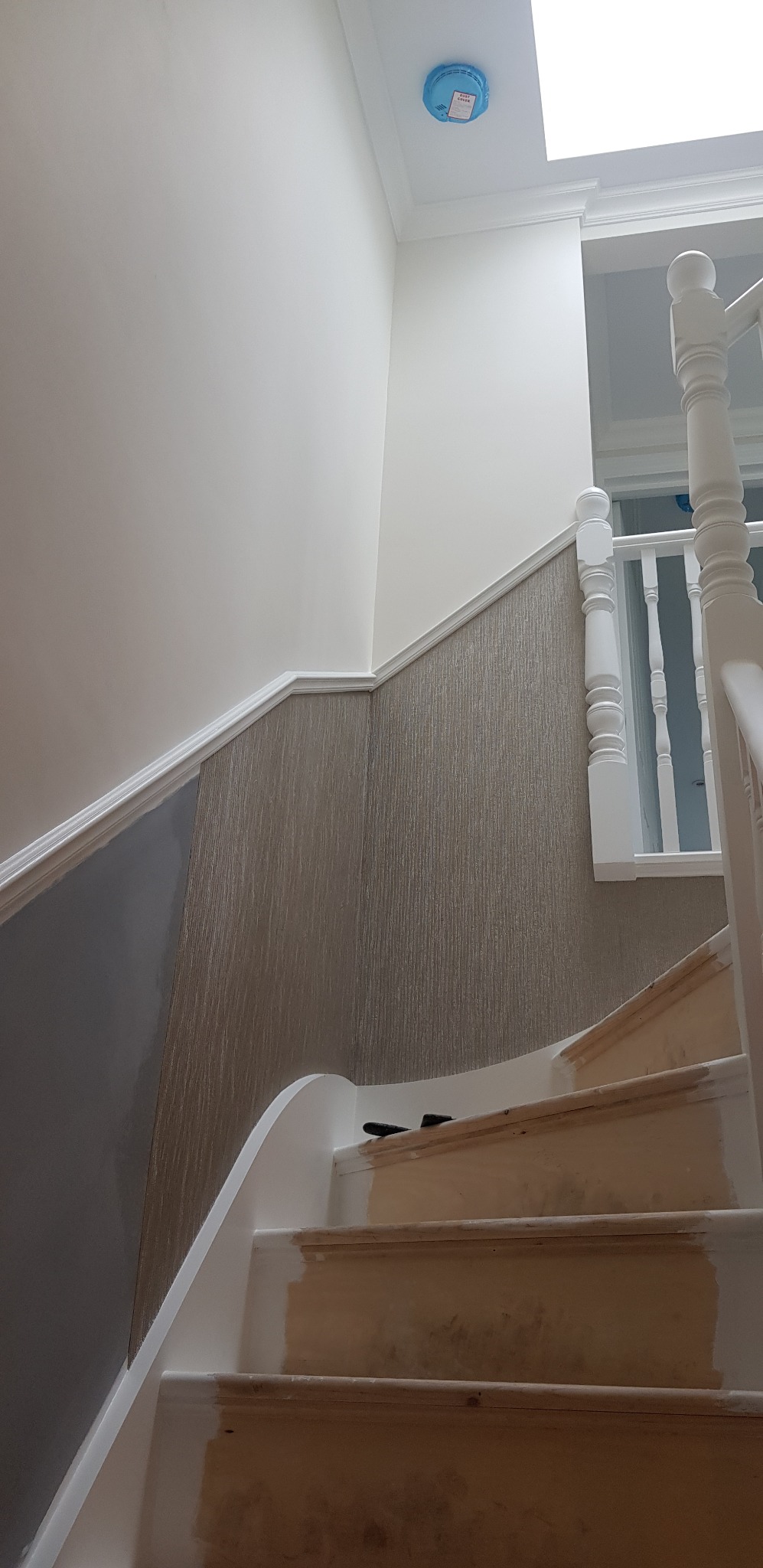 Schody w trakcie remontu: częściowo pomalowane stopnie, fragment szarej ściany, tapeta z delikatnym wzorem, biała balustrada i czujnik dymu na suficie.