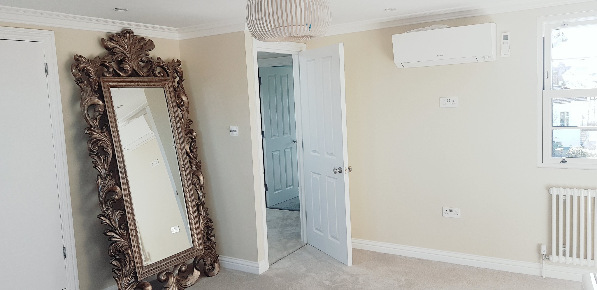 Jasne wnętrze pokoju po remoncie, ściany w odcieniach beżu, ozdobne lustro w złotej ramie, biała stolarka drzwiowa, klimatyzator na ścianie, grzejnik pod oknem.