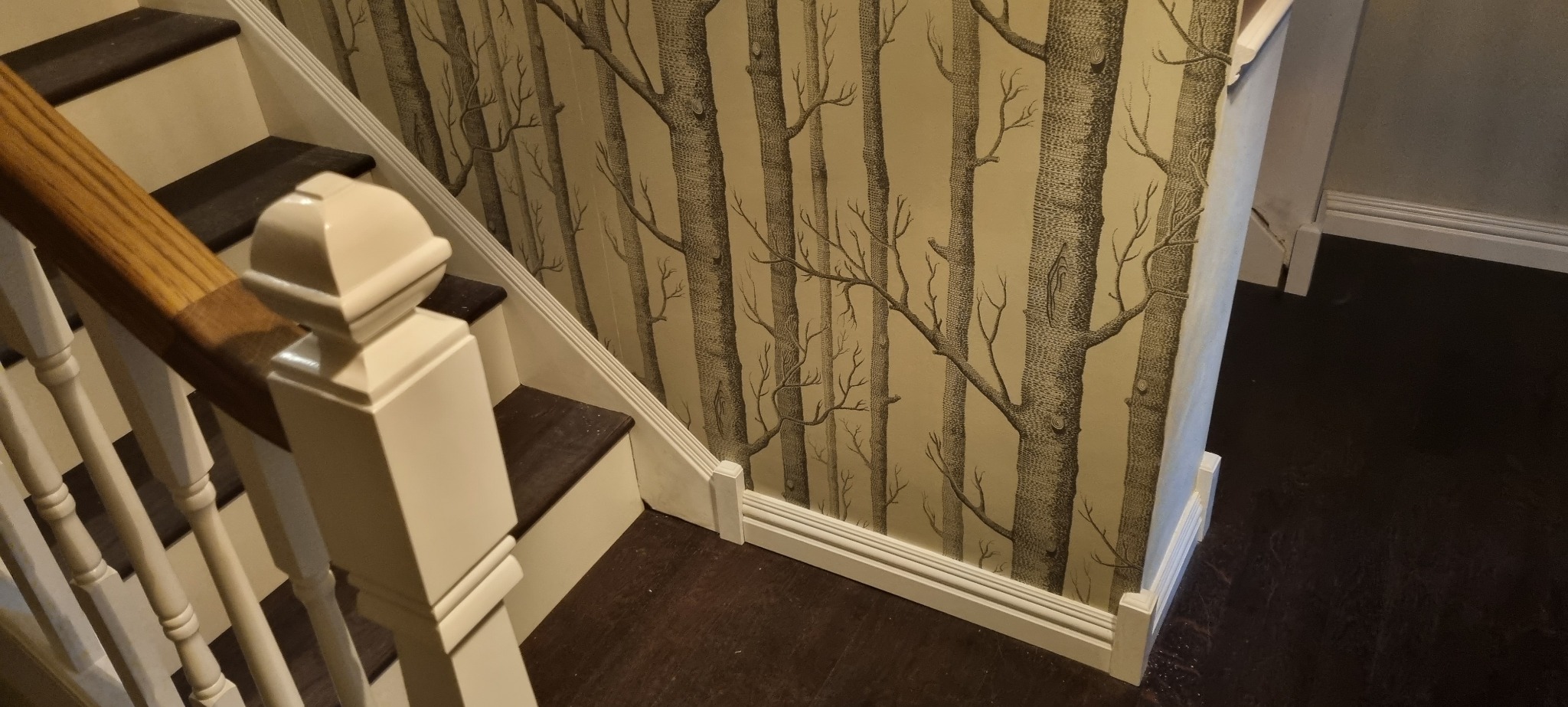 Klatka schodowa z tapetą w motywie drzew, drewniane schody z białymi balustradami i brązowymi stopniami.