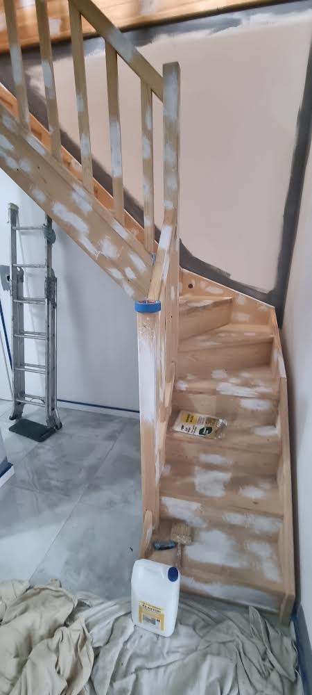 Drewniane schody z balustradą w trakcie zabezpieczania lub gruntowania, widoczne białe plamy farby, drabina aluminiowa w tle, wiadro farby i pędzel na zabezpieczonej folią podłodze.