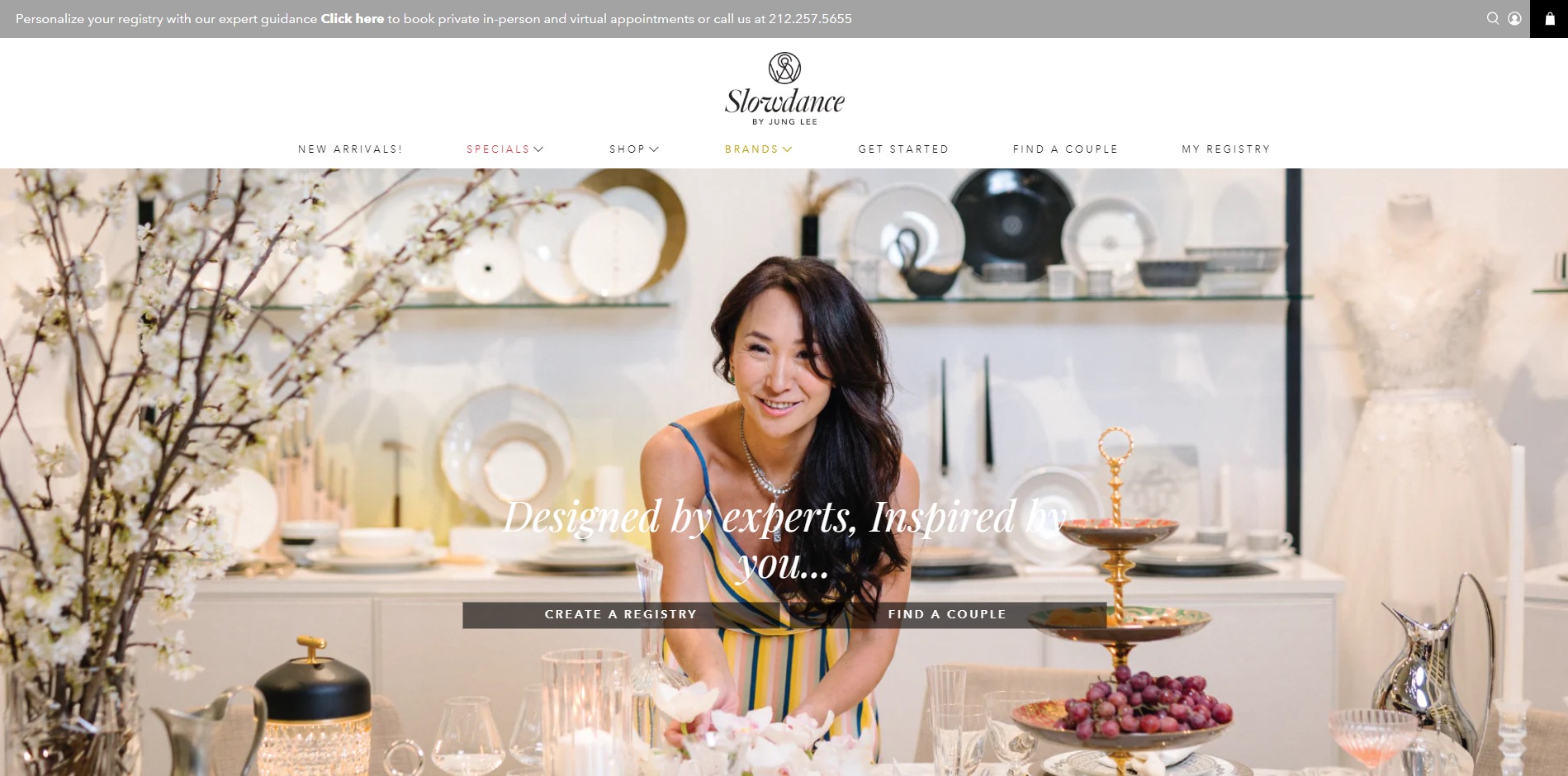 Kobieta w sukience w paski, uśmiechnięta, aranżuje kwiaty na stole z zastawą i dekoracjami, w tle półki z naczyniami i suknia ślubna.