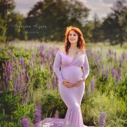 Magiczne Kadry - Fotografia Marzena Czura - Uśmiechnięta, ruda kobieta w liliowej sukni ciążowej, pozuje w plenerze wśród fioletowych kwiatów. Delikatne światło podkreśla naturalność sesji.