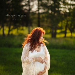 Magiczne Kadry - Fotografia Marzena Czura - Kobieta w ciąży, w zwiewnej koronkowej sukni, pozuje na tle zieleni. Rude włosy rozwiane na wietrze, delikatny uśmiech. Sesja w plenerze, naturalne światło.