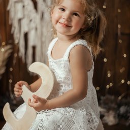 Magiczne Kadry - Fotografia Marzena Czura - Uśmiechnięta dziewczynka w białej sukience trzyma drewniany księżyc na tle dekoracji boho i światełek, uchwycona podczas sesji w Bochni.