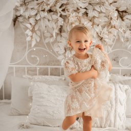 Magiczne Kadry - Fotografia Marzena Czura - Uśmiechnięta blond dziewczynka w koronkowej sukience stoi na białym łóżku z dekoracją kwiatową, trzymając fragment materiału. Jasne, naturalne światło.