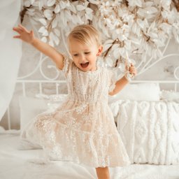 Magiczne Kadry - Fotografia Marzena Czura - Radosna blondwłosa dziewczynka w koronkowej sukience tańczy na białym łóżku z dekoracją kwiatową w tle. Uśmiech, ruch, jasne wnętrze, naturalne światło.