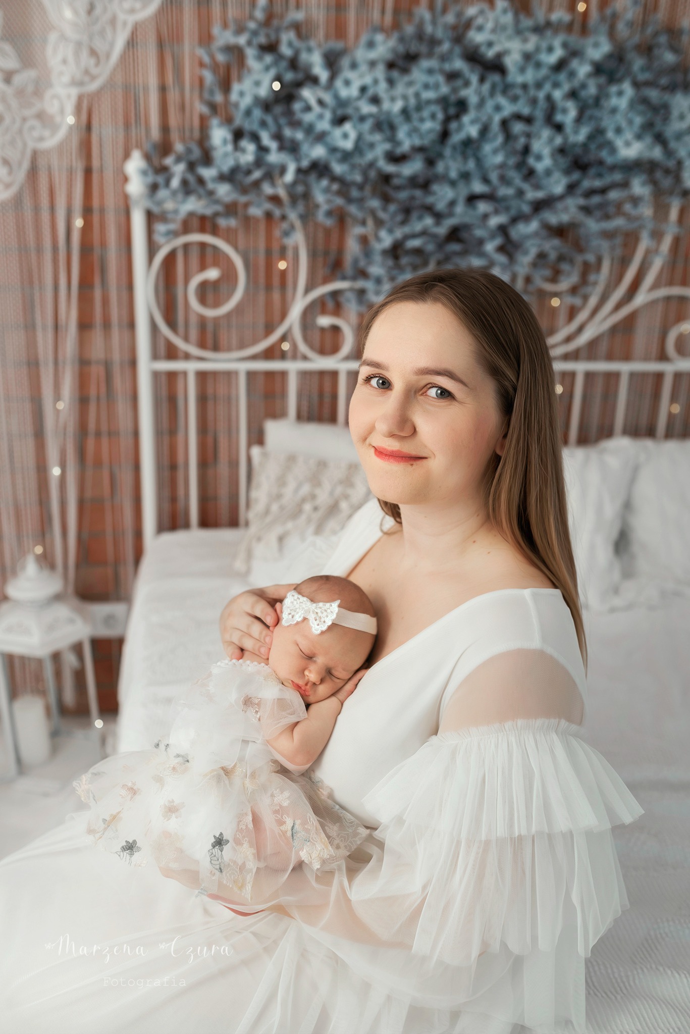 Portret matki w białej sukni z falbanami trzymającej śpiące niemowlę w tiulowej sukience z aplikacjami kwiatowymi i kokardą na głowie, na tle łóżka z dekoracjami.