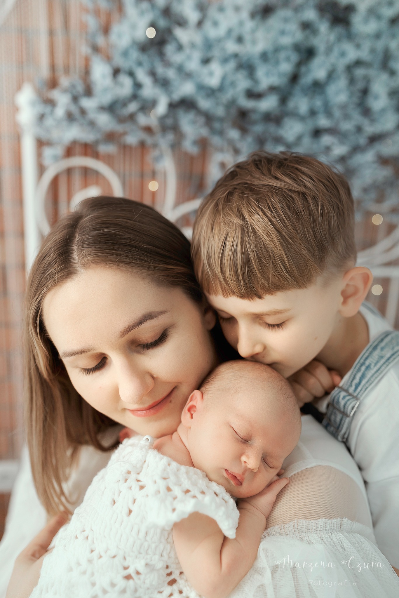 Mama trzyma śpiącego noworodka w białym ażurowym kocyku, starszy brat całuje dziecko w główkę, w tle dekoracja z białych gałązek.