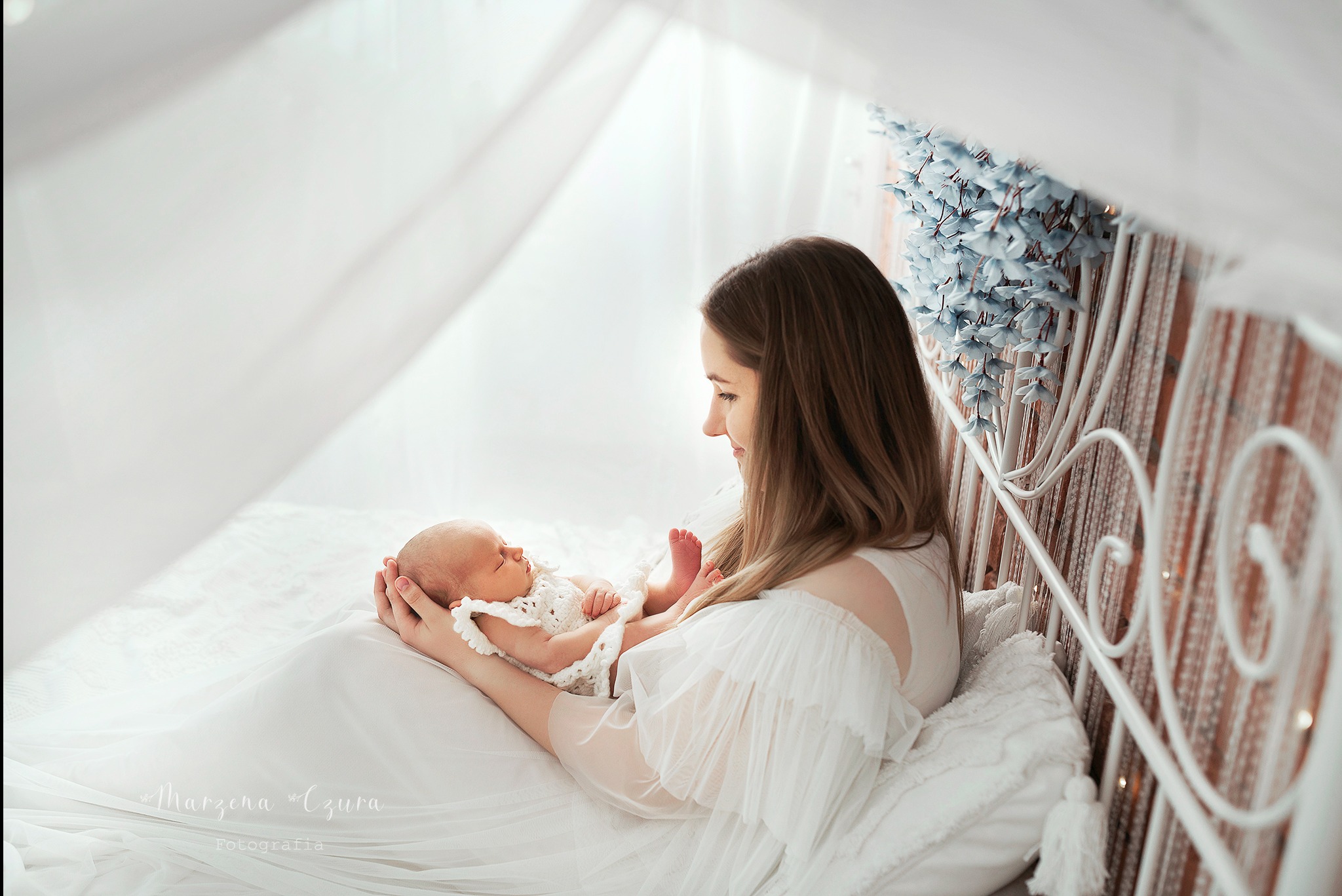 Matka w białej sukni trzyma na kolanach śpiącego noworodka w białym ubranku na tle białego łóżka z dekoracjami kwiatowymi.