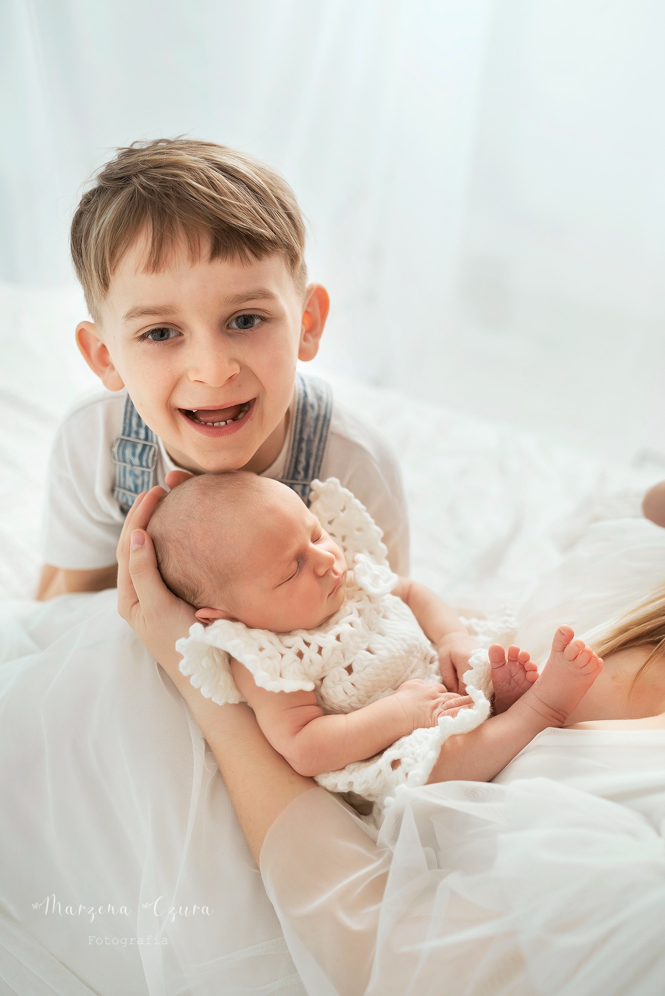 Starszy brat uśmiecha się, opierając głowę na główce śpiącego noworodka ubranego w białą, dzierganą sukienkę, trzymanego przez matkę.
