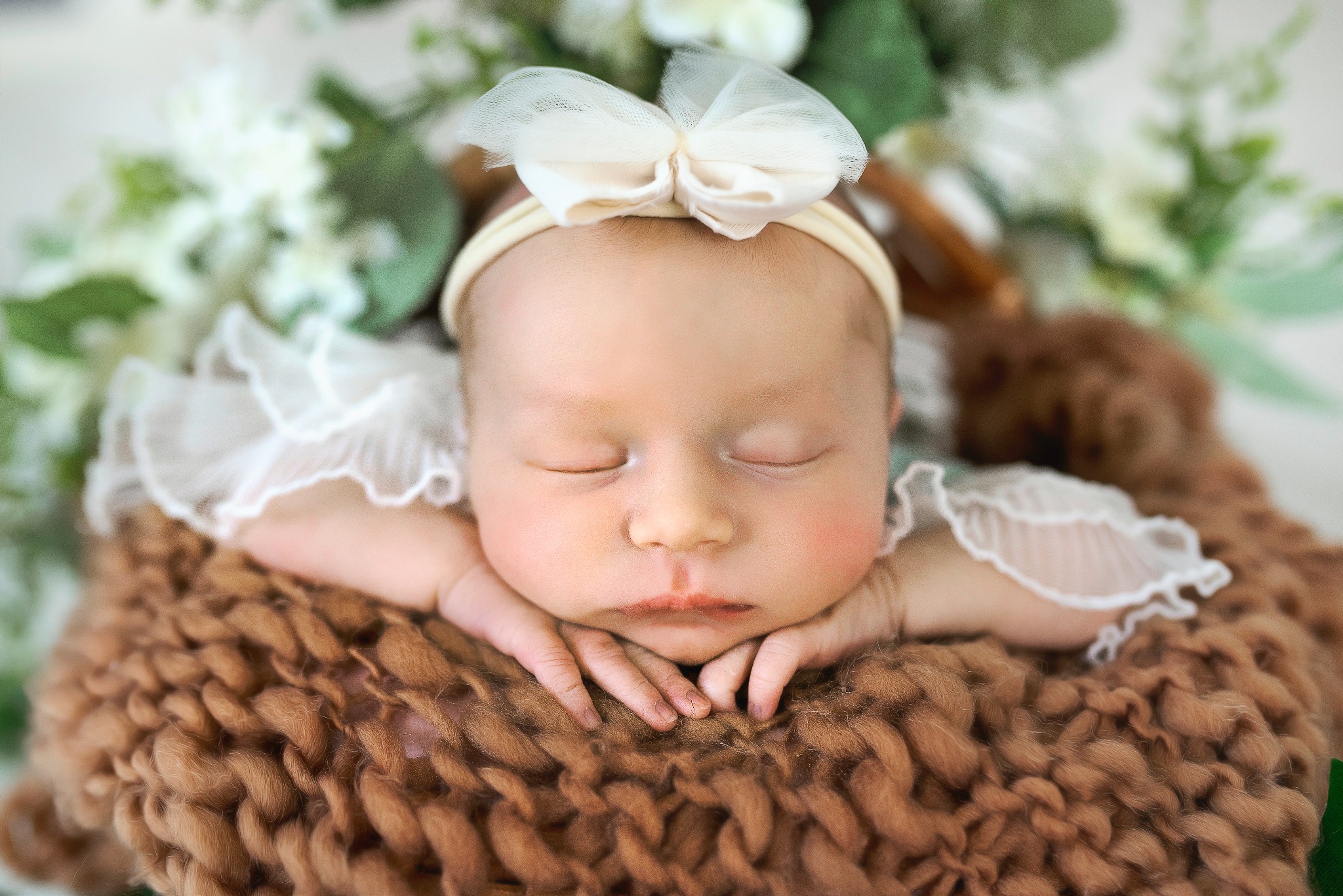 Śpiąca noworodka z zamkniętymi oczami, w białej opasce z kokardką, ułożona w brązowym, dzierganym koszyku na tle białych kwiatów.