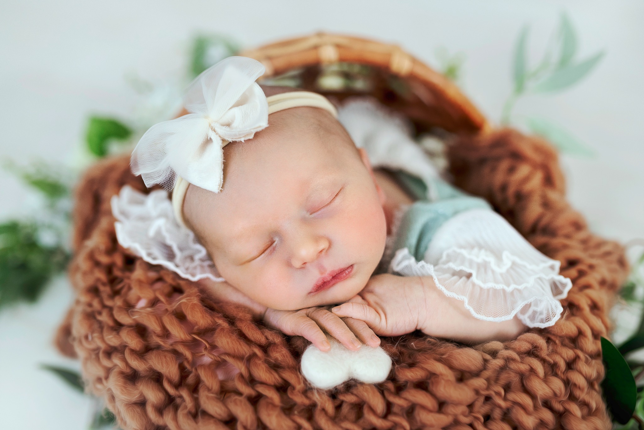 Śpiący noworodek z białą kokardą na głowie, ułożony w brązowym, dzierganym koszyku z koronkowym wykończeniem i zielonymi akcentami roślinnymi w tle.