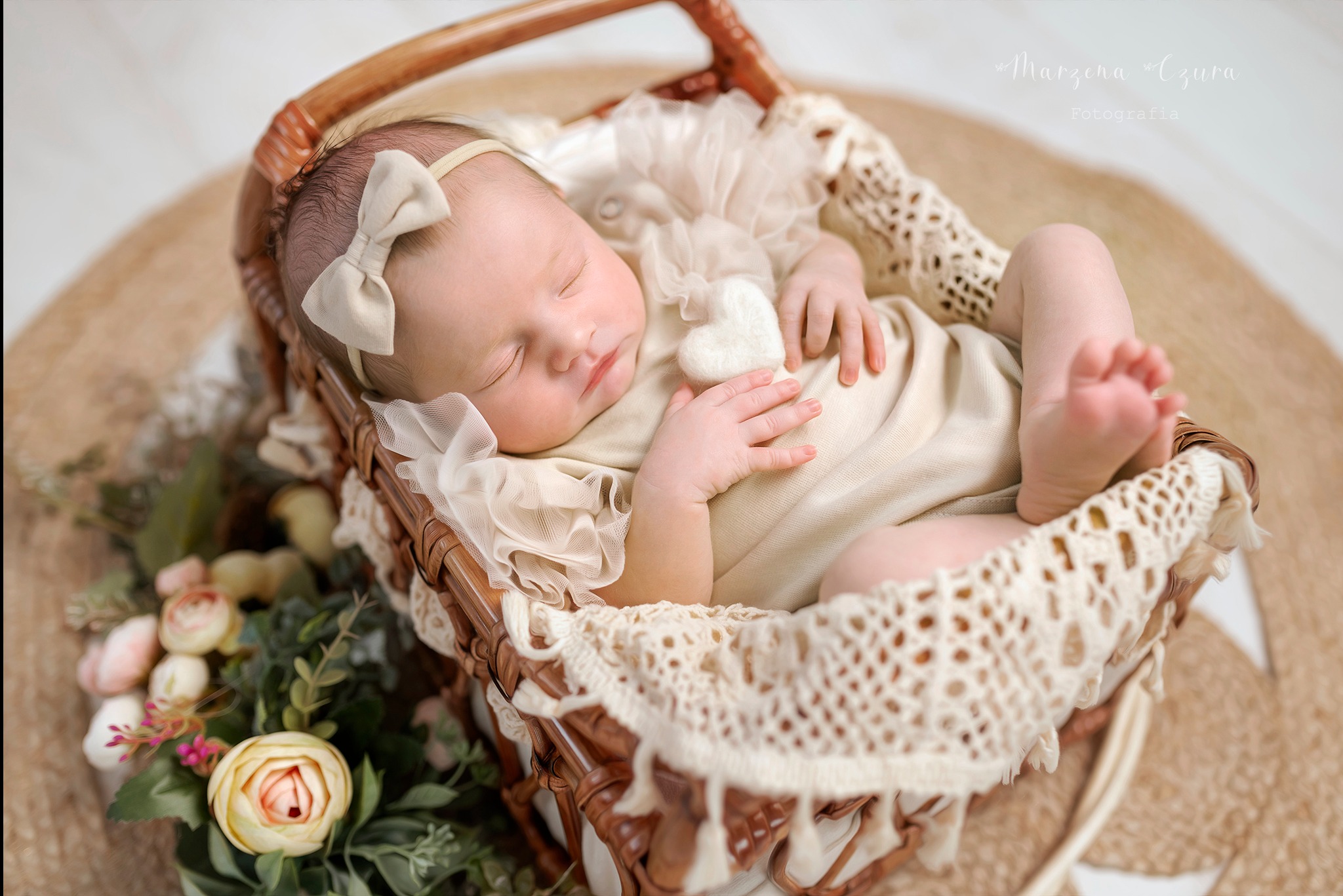 Śpiący noworodek z kokardką na głowie, ułożony w wiklinowym koszu wyściełanym koronką i kwiatami, sfotografowany w Bochni.
