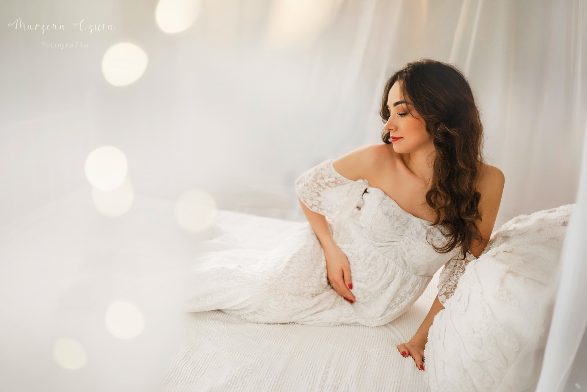 Kobieta w koronkowej sukni, w delikatnej sesji ciążowej w studio, z miękkim światłem i subtelnym tłem, leżąca na białym łóżku. Romantyczny nastrój.