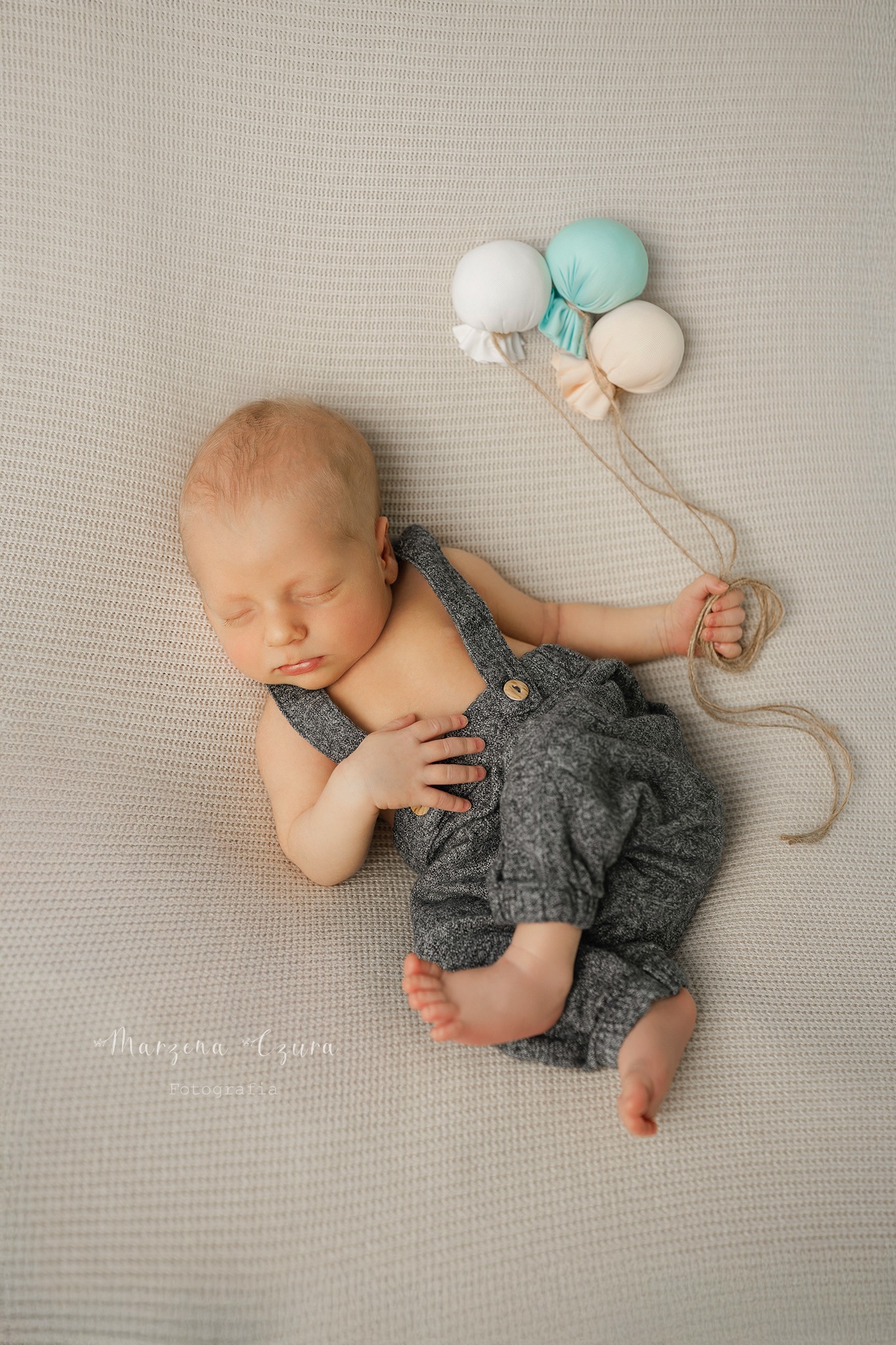 Śpiący noworodek w szarym ubranku leży na beżowym tle, trzymając w rączce sznurki od trzech pastelowych balonów. Delikatne światło podkreśla detale.