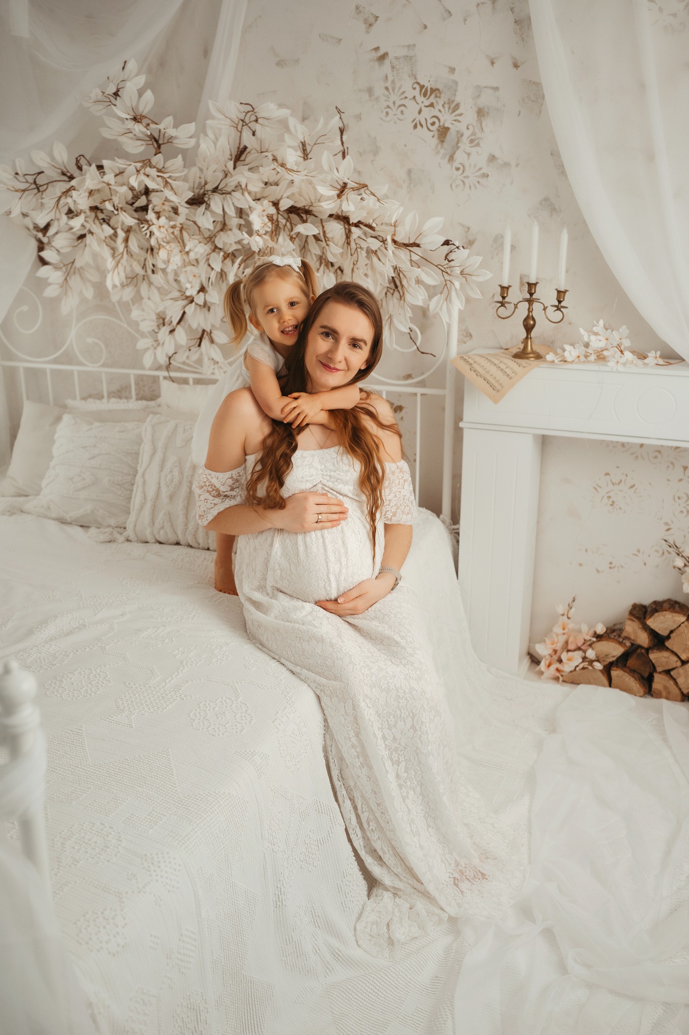 Ciąża w bieli: Uśmiechnięta przyszła mama w koronkowej sukni, siedzi na łóżku z córeczką w jasnym pokoju, dekorowanym kwiatami i kominkiem.