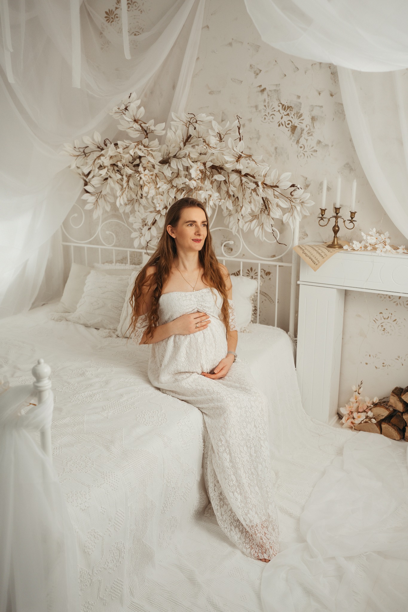 Kobieta w ciąży w białej sukni siedzi na łóżku w jasnym pokoju z dekoracjami kwiatowymi i kominkiem. Romantyczna sesja w stylu vintage.