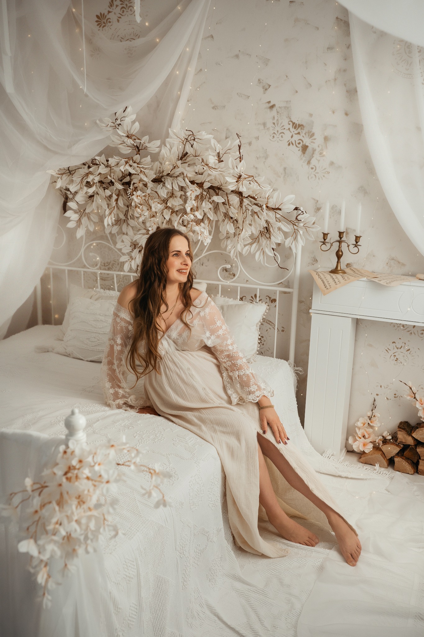 Uśmiechnięta kobieta w zwiewnej sukni siedzi na białym łóżku w dekoracyjnej scenerii z kwiatami i świecami, pozując do sesji ciążowej w jasnym, naturalnym świetle.