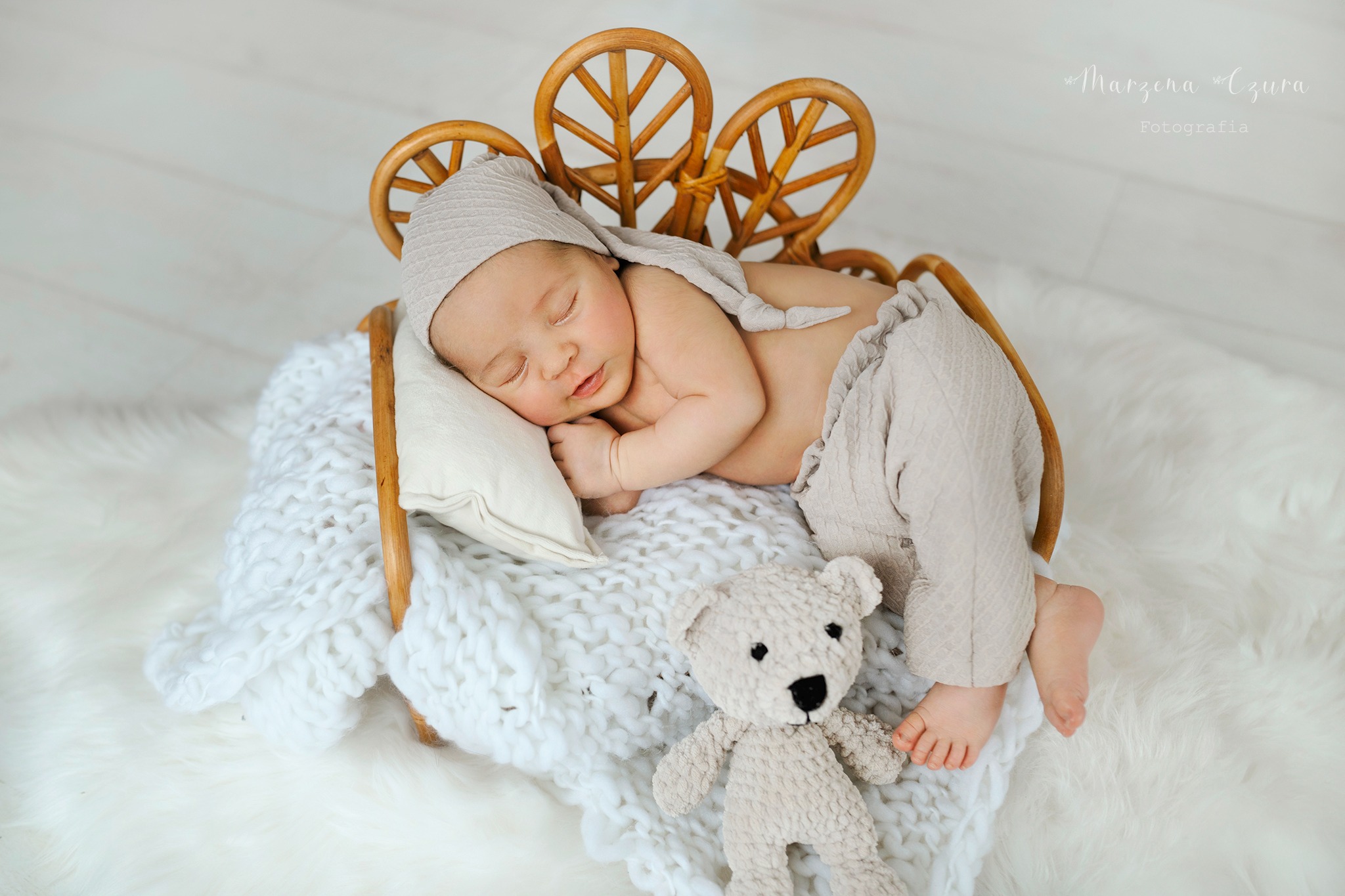 Śpiący noworodek w beżowym ubranku i czapce, leżący na białym kocyku w wiklinowym łóżeczku z pluszowym misiem. Delikatne, naturalne światło.