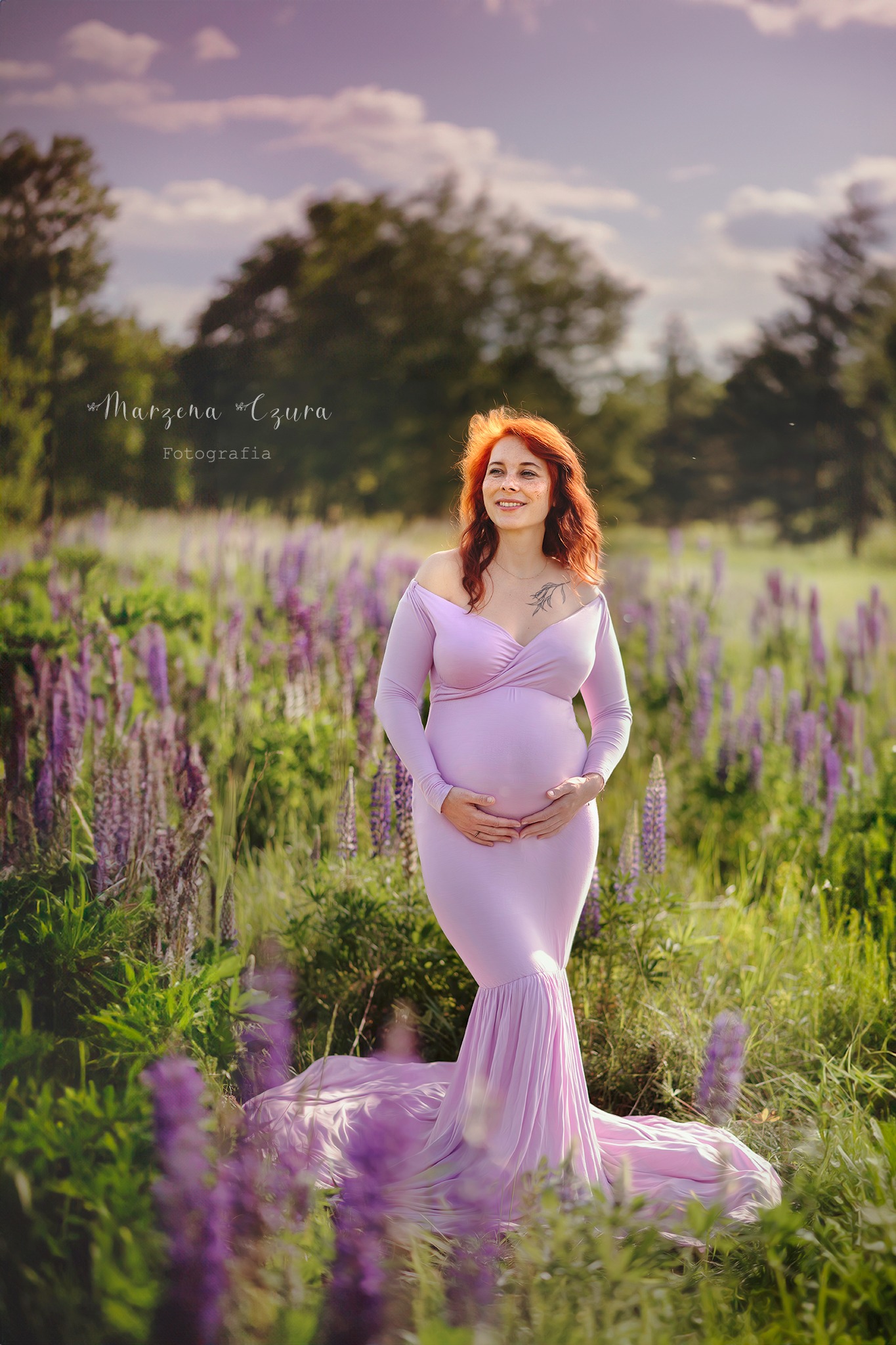 Uśmiechnięta, ruda kobieta w liliowej sukni ciążowej, pozuje w plenerze wśród fioletowych kwiatów. Delikatne światło podkreśla naturalność sesji.