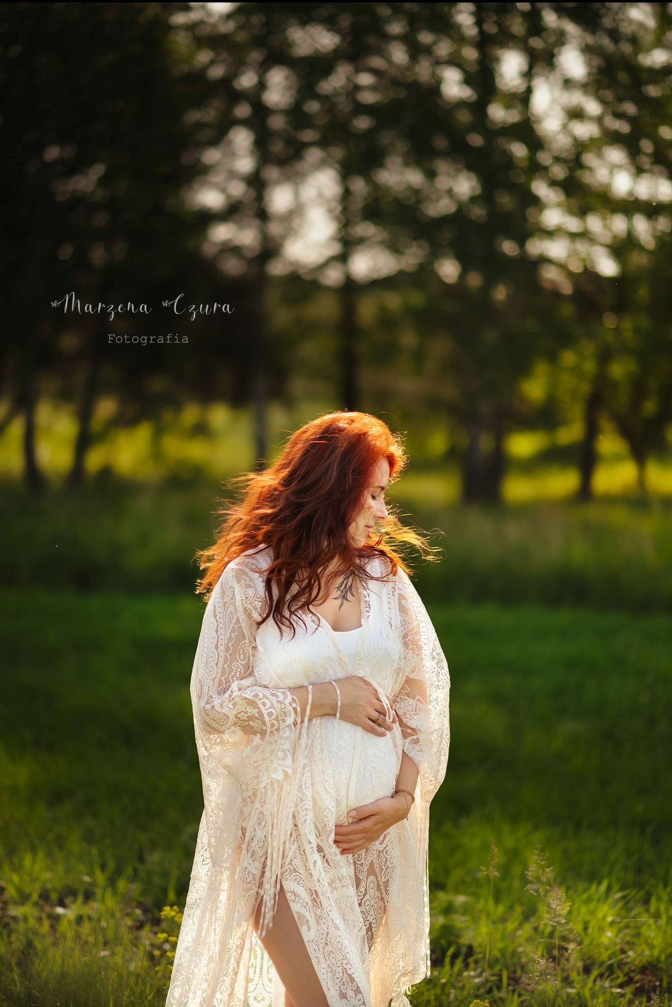 Kobieta w ciąży, w zwiewnej koronkowej sukni, pozuje na tle zieleni. Rude włosy rozwiane na wietrze, delikatny uśmiech. Sesja w plenerze, naturalne światło.