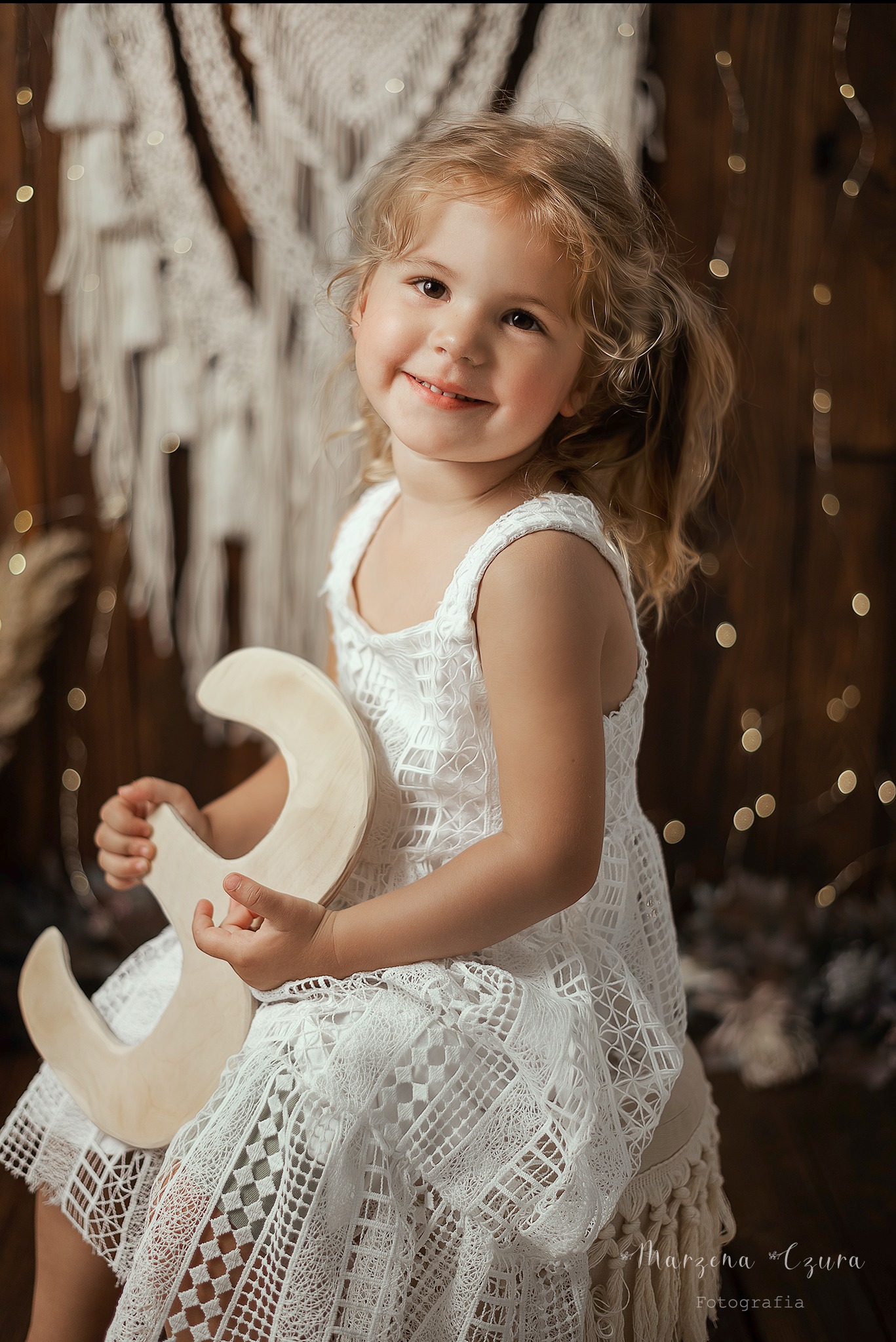 Uśmiechnięta dziewczynka w białej sukience trzyma drewniany księżyc na tle dekoracji boho i światełek, uchwycona podczas sesji w Bochni.