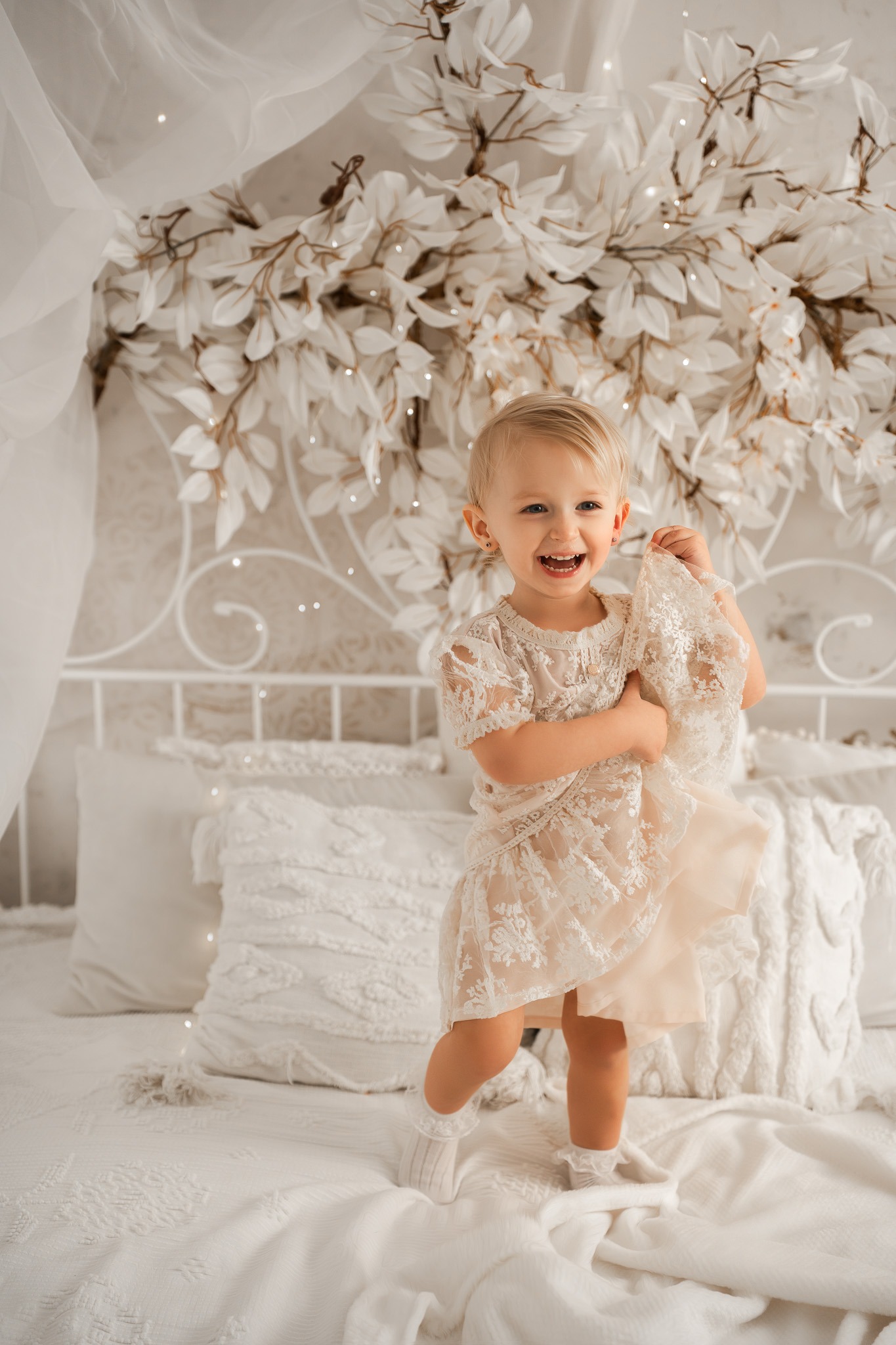 Uśmiechnięta blond dziewczynka w koronkowej sukience stoi na białym łóżku z dekoracją kwiatową, trzymając fragment materiału. Jasne, naturalne światło.