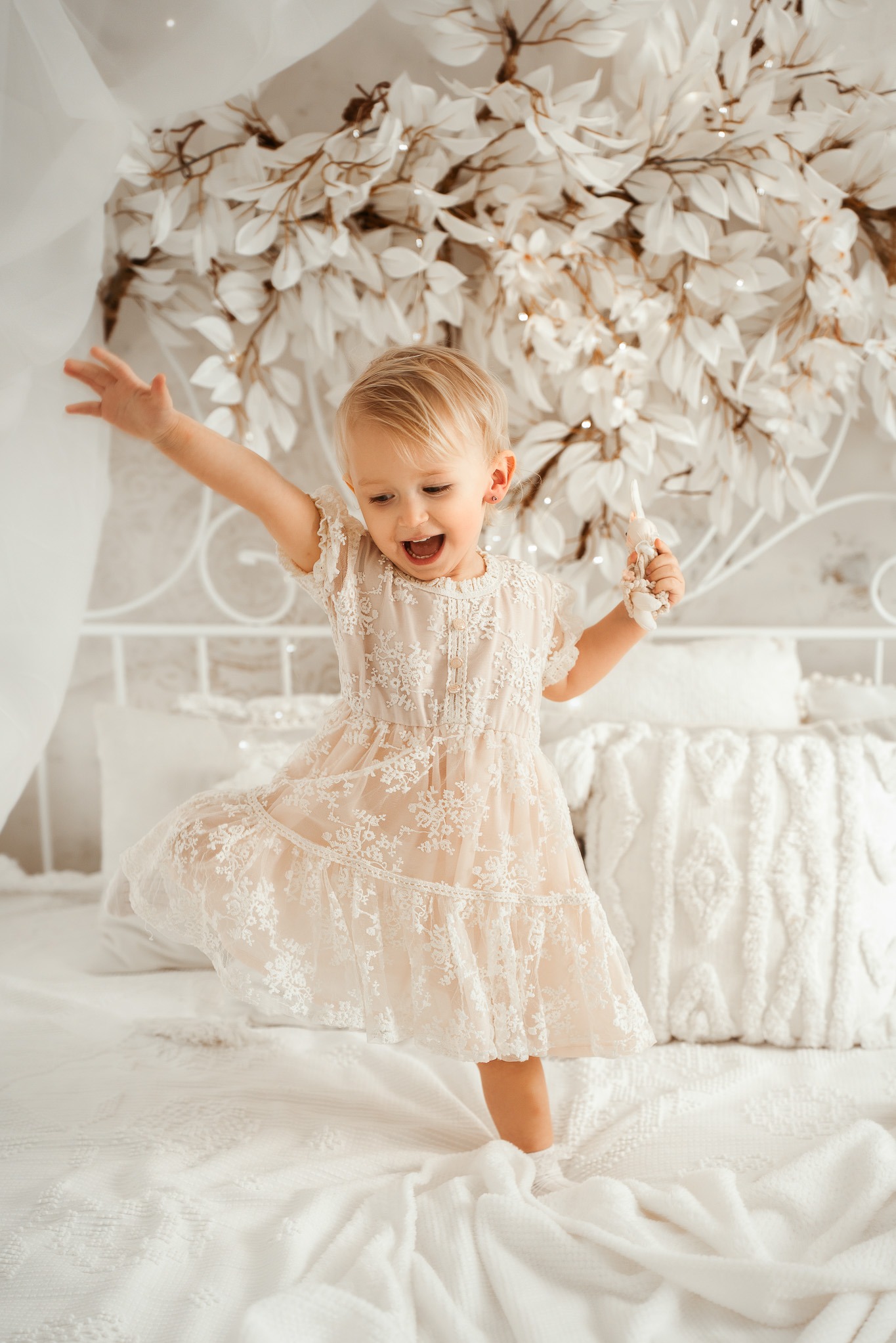 Radosna blondwłosa dziewczynka w koronkowej sukience tańczy na białym łóżku z dekoracją kwiatową w tle. Uśmiech, ruch, jasne wnętrze, naturalne światło.