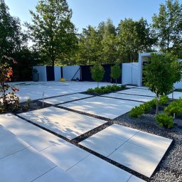 Szymon w Ogrodzie - Nowoczesna aranżacja ogrodu z jasnymi płytami chodnikowymi, czarnym grysem i młodymi drzewkami. Minimalistyczny design z wyraźnymi liniami i geometrycznym układem.