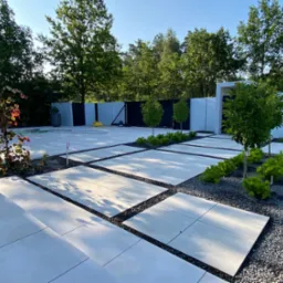 Nowoczesna aranżacja ogrodu z jasnymi płytami chodnikowymi, czarnym grysem i młodymi drzewkami. Minimalistyczny design z wyraźnymi liniami i geometrycznym układem.