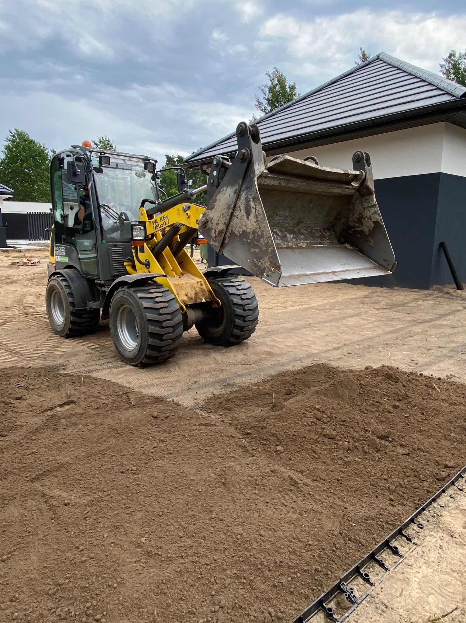 Żółto-szara ładowarka na przygotowanym terenie pod trawnik, z widocznymi obrzeżami. W tle dom z szarą dachówką i dwukolorową elewacją.