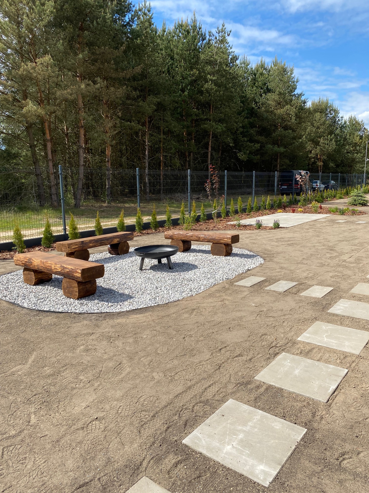 Ogród z drewnianymi ławkami wokół paleniska na żwirowym podłożu. Ścieżka z płyt prowadzi przez teren, w tle drzewa i ogrodzenie.
