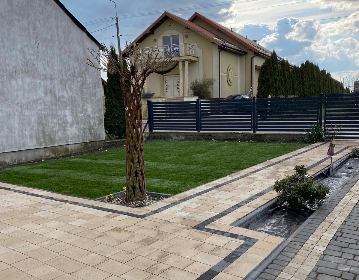 Zadbany trawnik z kostką brukową i ozdobnym drzewem plecionym na tle domu z balkonem i ogrodzeniem. Widoczne elementy małej architektury ogrodowej.