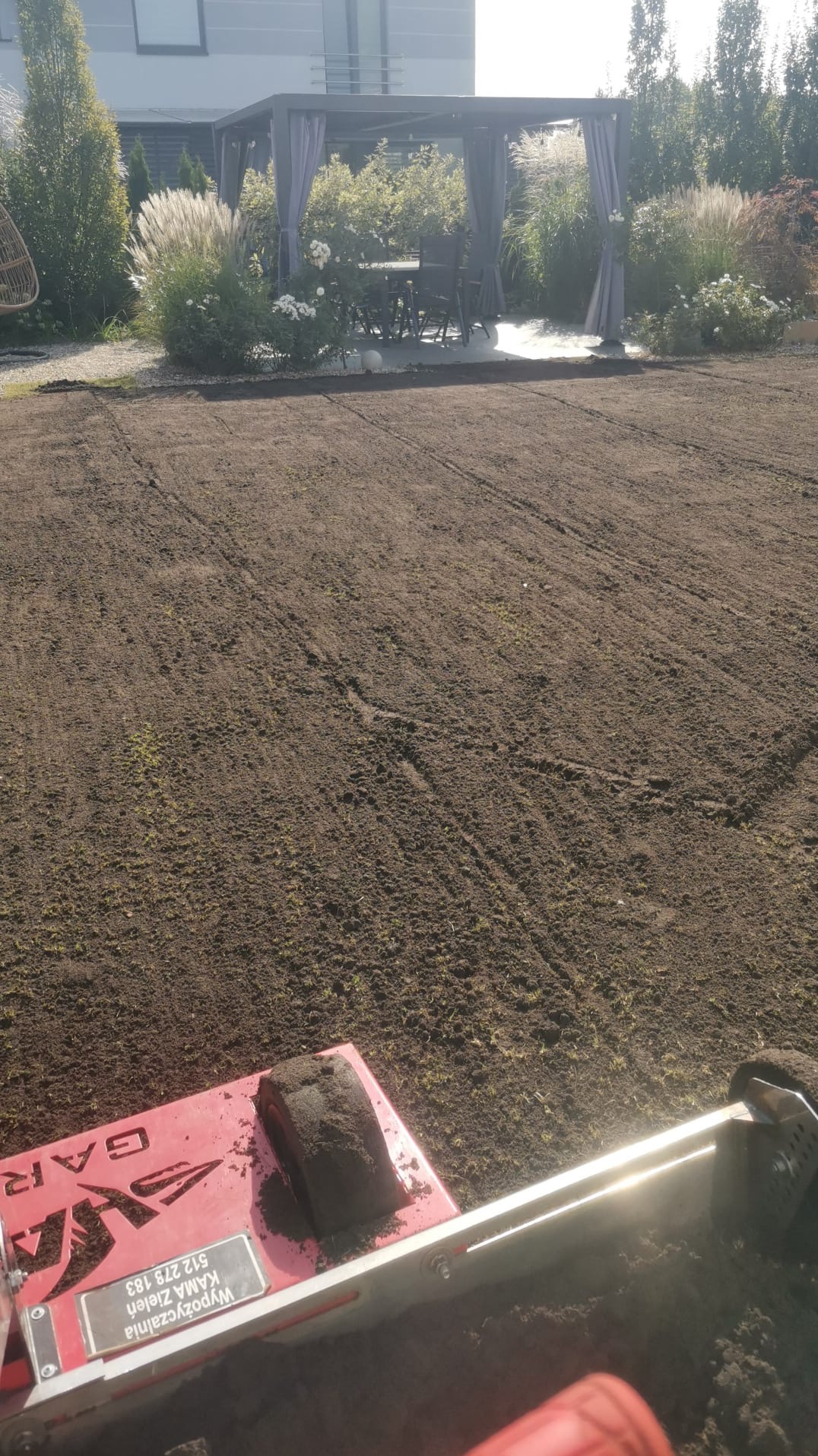 Przygotowany grunt pod trawnik z widocznym urządzeniem do wyrównywania, w tle pergola i ozdobne trawy. Układanie trawy z rolki.