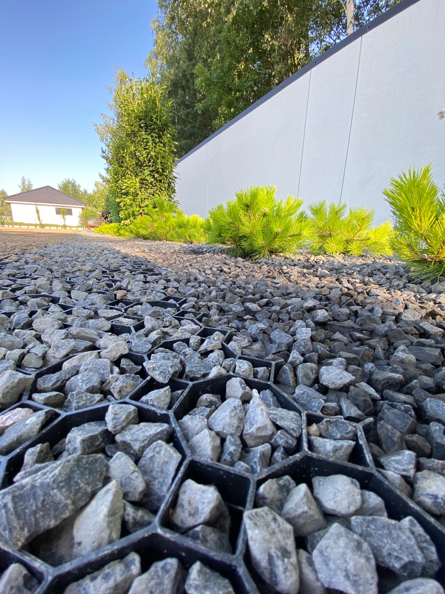 Układanie kamieni na geokracie w ogrodzie, niskie ujęcie podkreśla perspektywę, w tle rząd małych sosen i biały mur.
