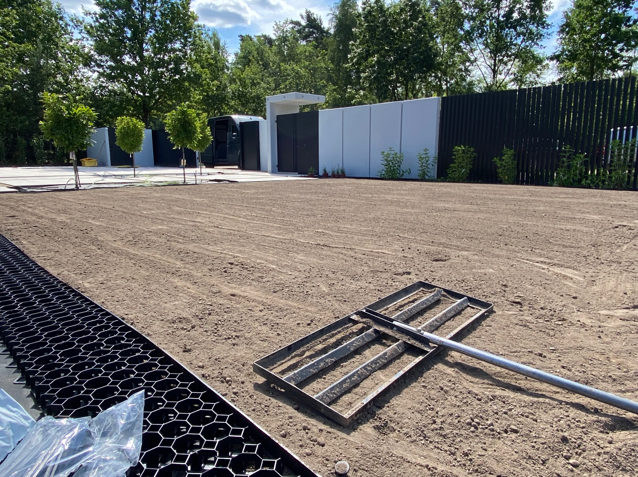 Przygotowane podłoże pod trawnik z rolki, wyrównane grabiami. W tle małe drzewka, ogrodzenie z paneli i budynek gospodarczy. Elementy drenażowe w lewym dolnym rogu.