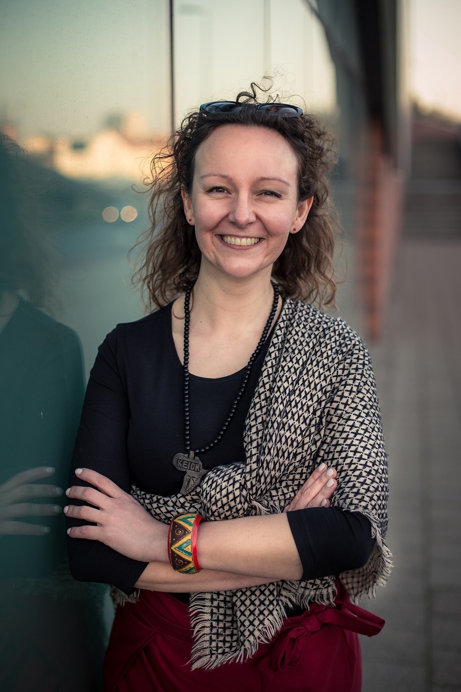 Uśmiechnięta kobieta z kręconymi włosami, w czarnej bluzce i wzorzystej narzutce, stoi na zewnątrz z założonymi rękami, prezentując etniczną biżuterię i bransoletkę.