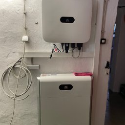 NOVA GREEN - Biały falownik Huawei i moduł magazynowania energii zamontowane na pomalowanej na biało ścianie z widocznymi przewodami i elementami instalacji elektrycznej.