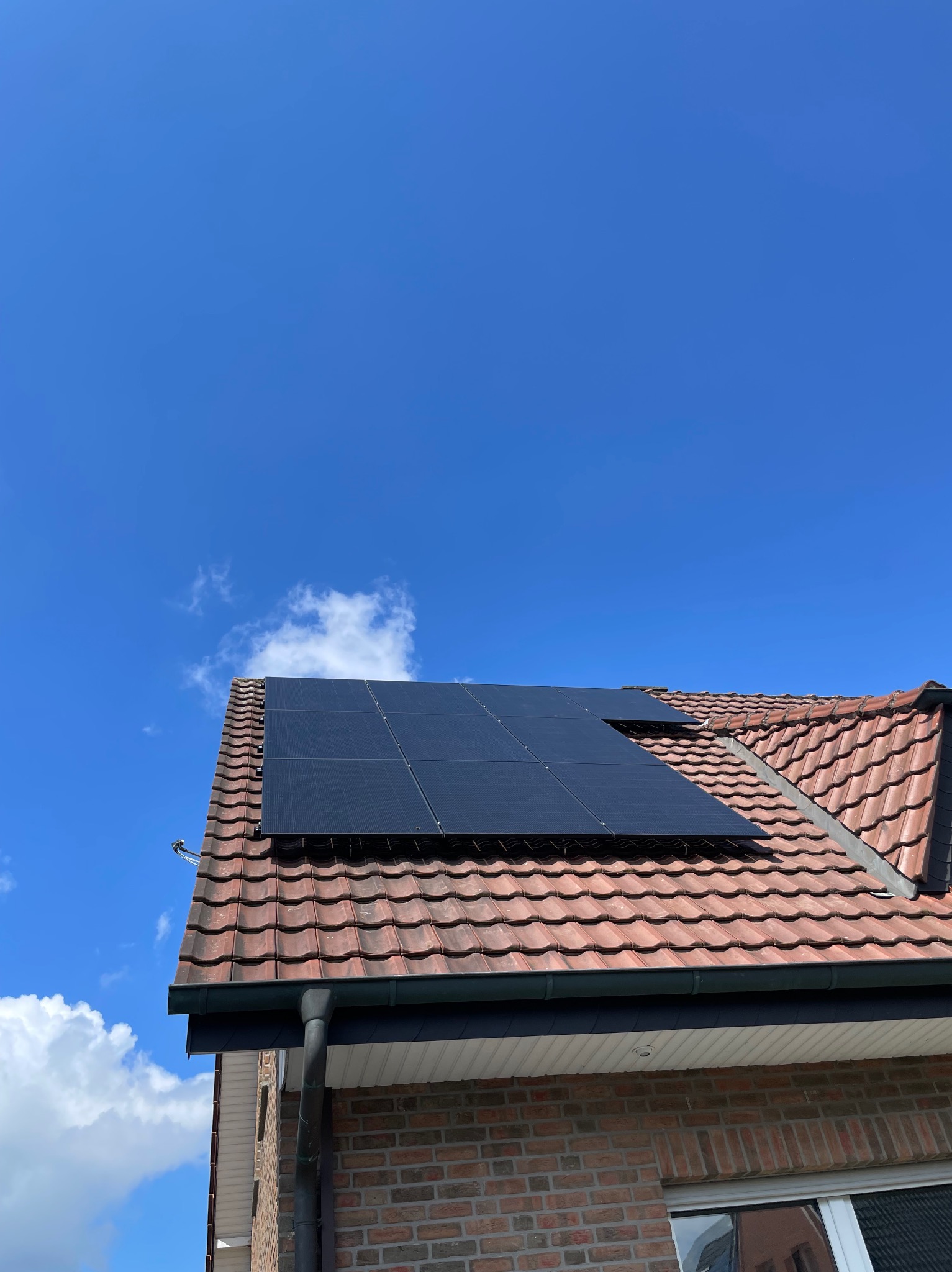 Częściowy widok paneli słonecznych na czerwonym dachu z dachówki ceramicznej, na tle błękitnego nieba z nielicznymi obłokami, widoczny fragment rynny i ściany budynku z cegły.