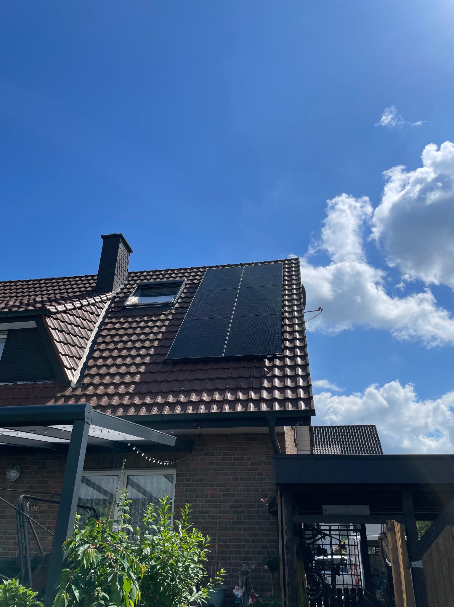 Panele słoneczne na brązowym, spadzistym dachu domu jednorodzinnego pod błękitnym niebem z białymi chmurami. Widoczny komin i okno dachowe.