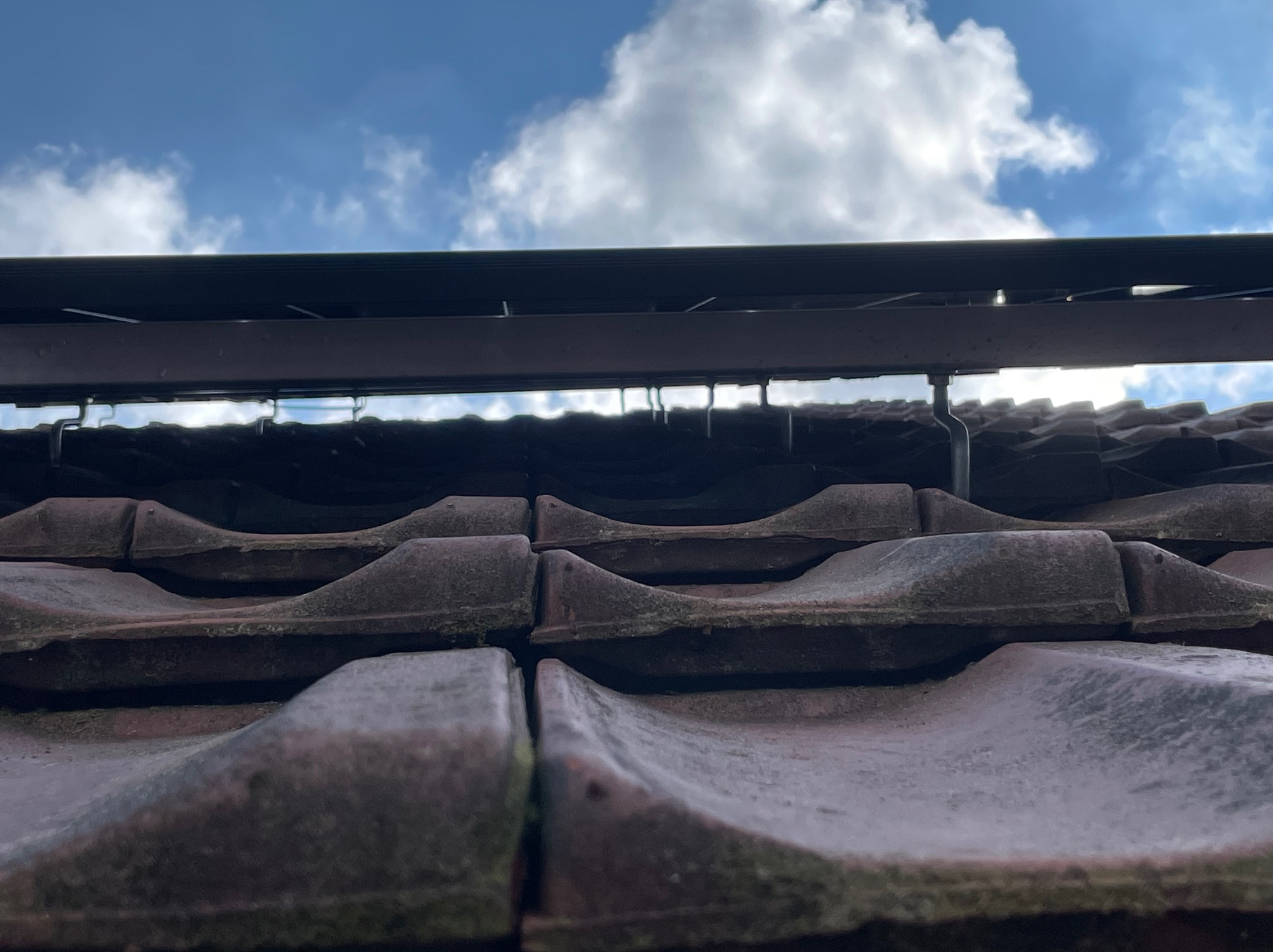 Montaż konstrukcji wsporczej paneli fotowoltaicznych na dachu krytym starą, brudną dachówką ceramiczną, widok z dołu na błękitne niebo z chmurami.