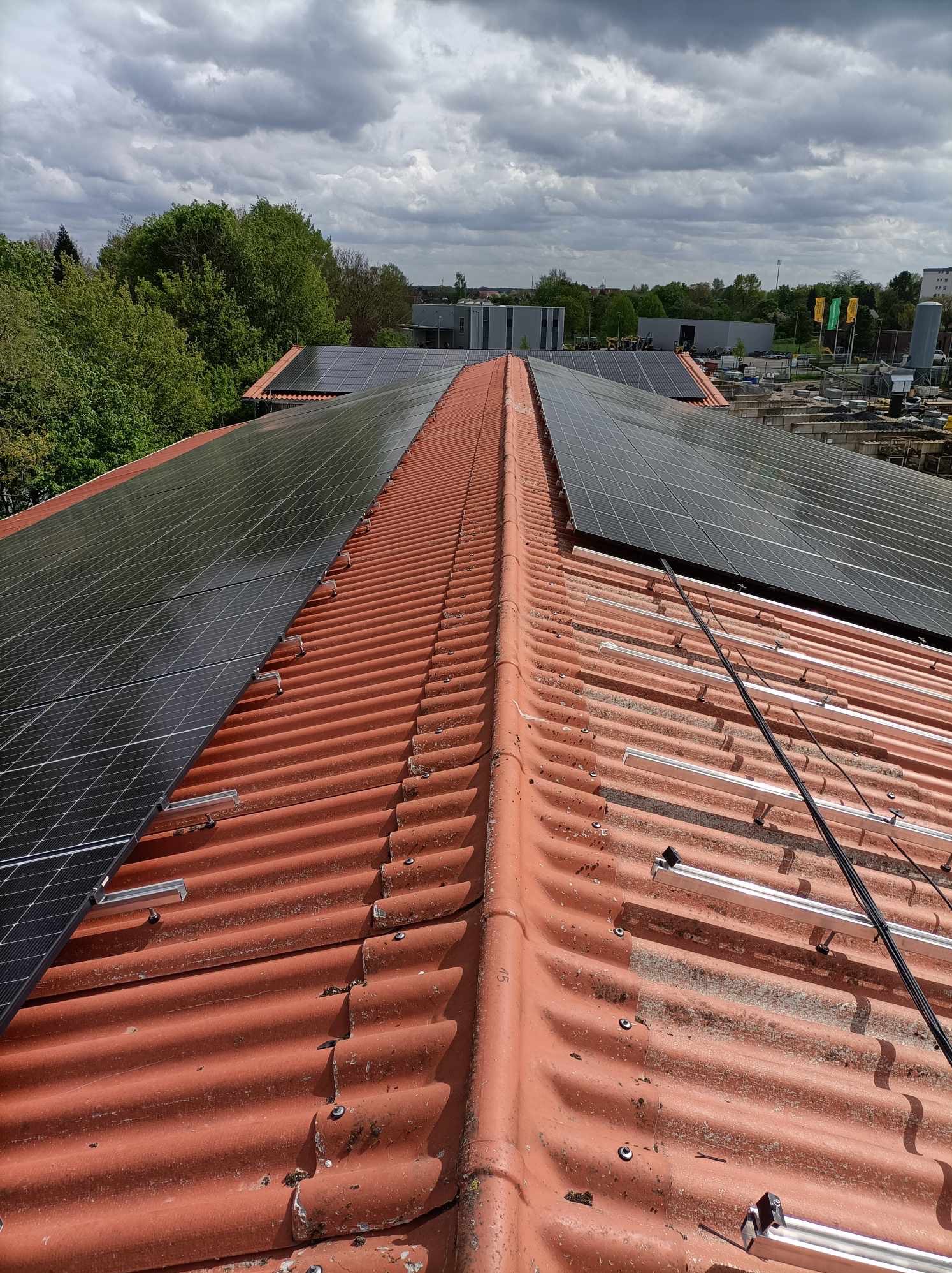 Częściowo zainstalowane panele na falistej, czerwonej dachówce. Widoczne aluminiowe szyny montażowe i kable. W tle drzewa i budynki pod pochmurnym niebem.