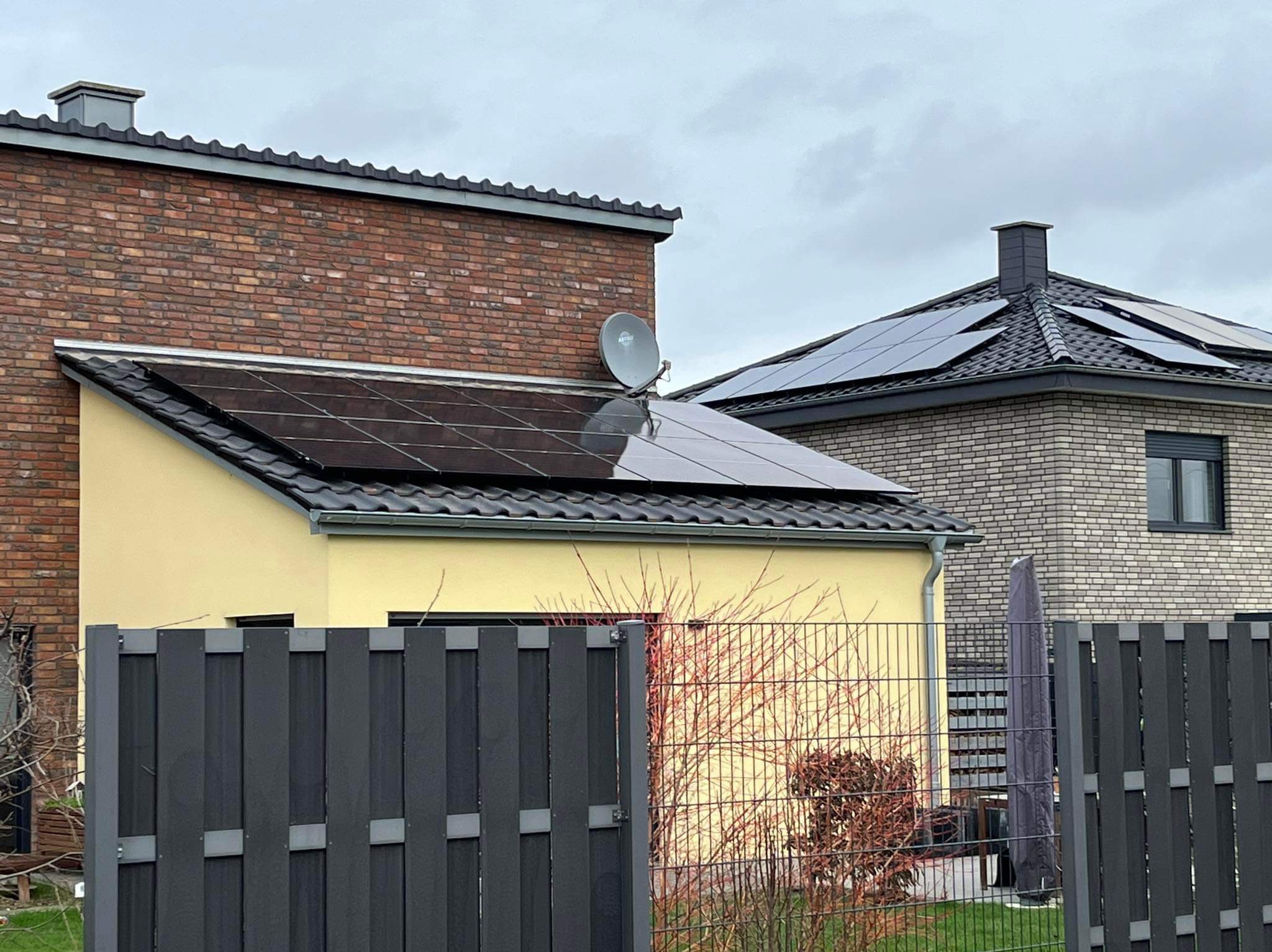 Dwa domy z panelami słonecznymi na dachach, widoczne zza grafitowego ogrodzenia. Na dachu żółtego domu antena satelitarna.