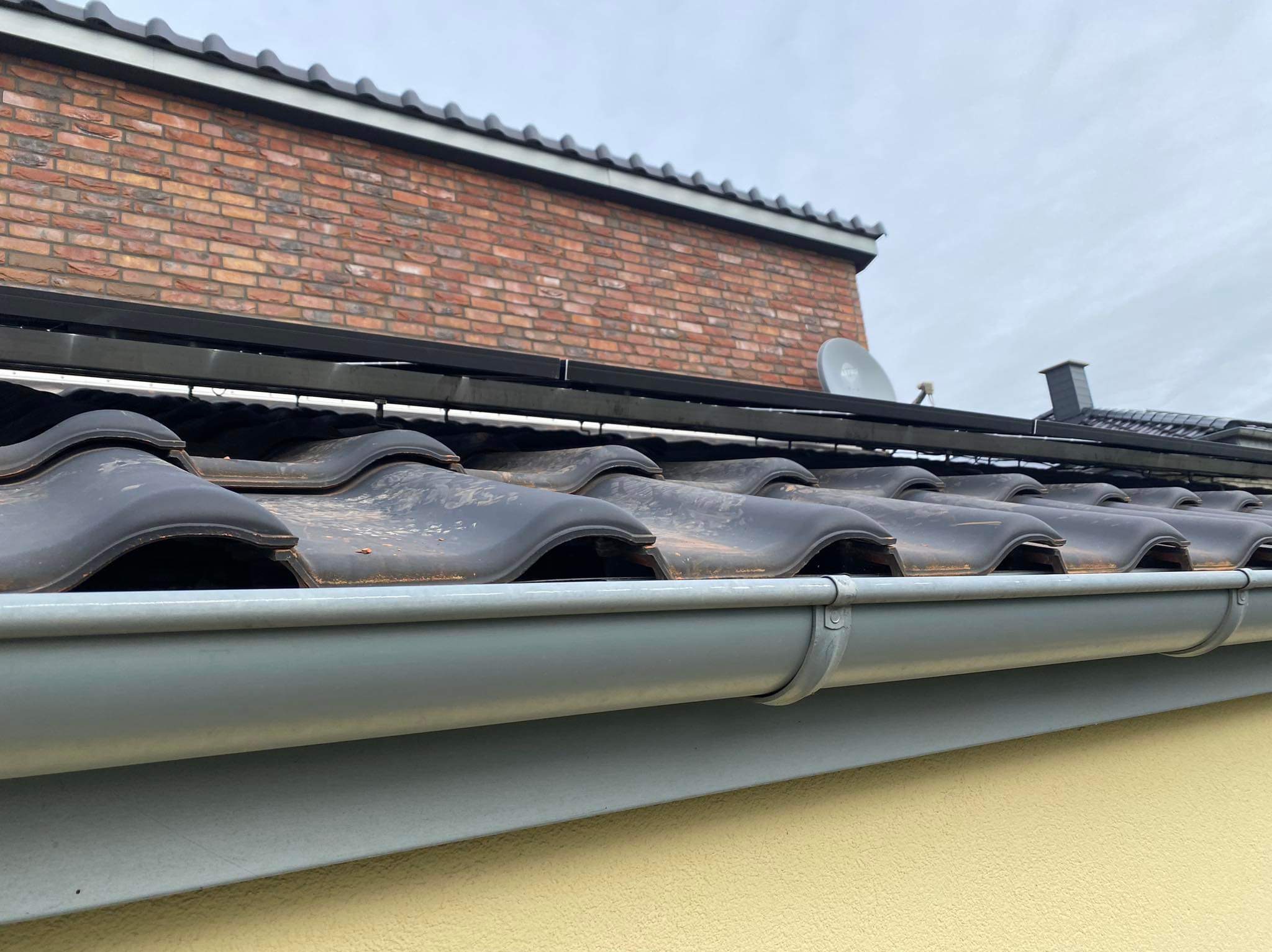 Ciemna, ceramiczna dachówka z szarą rynną i fragmentem ściany z cegły w tle, widok z dołu.