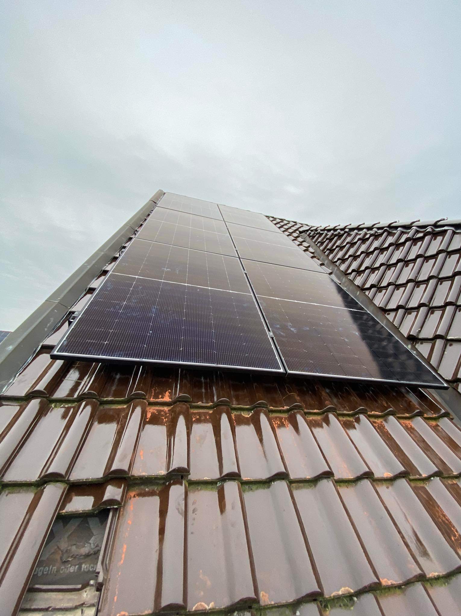 Panele słoneczne zamontowane na brązowym dachu z dachówki ceramicznej, widok z dołu, pochmurne niebo w tle.