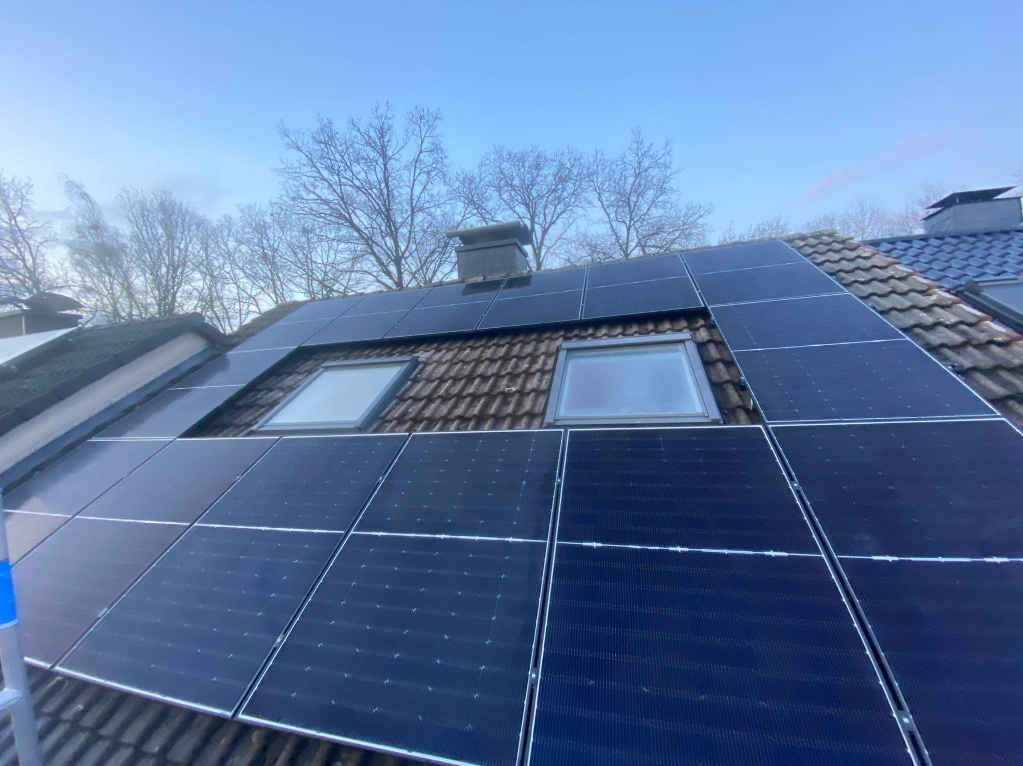 Panele fotowoltaiczne zamontowane na skośnym dachu z dachówką ceramiczną, widoczne okna dachowe i komin na tle błękitnego nieba.