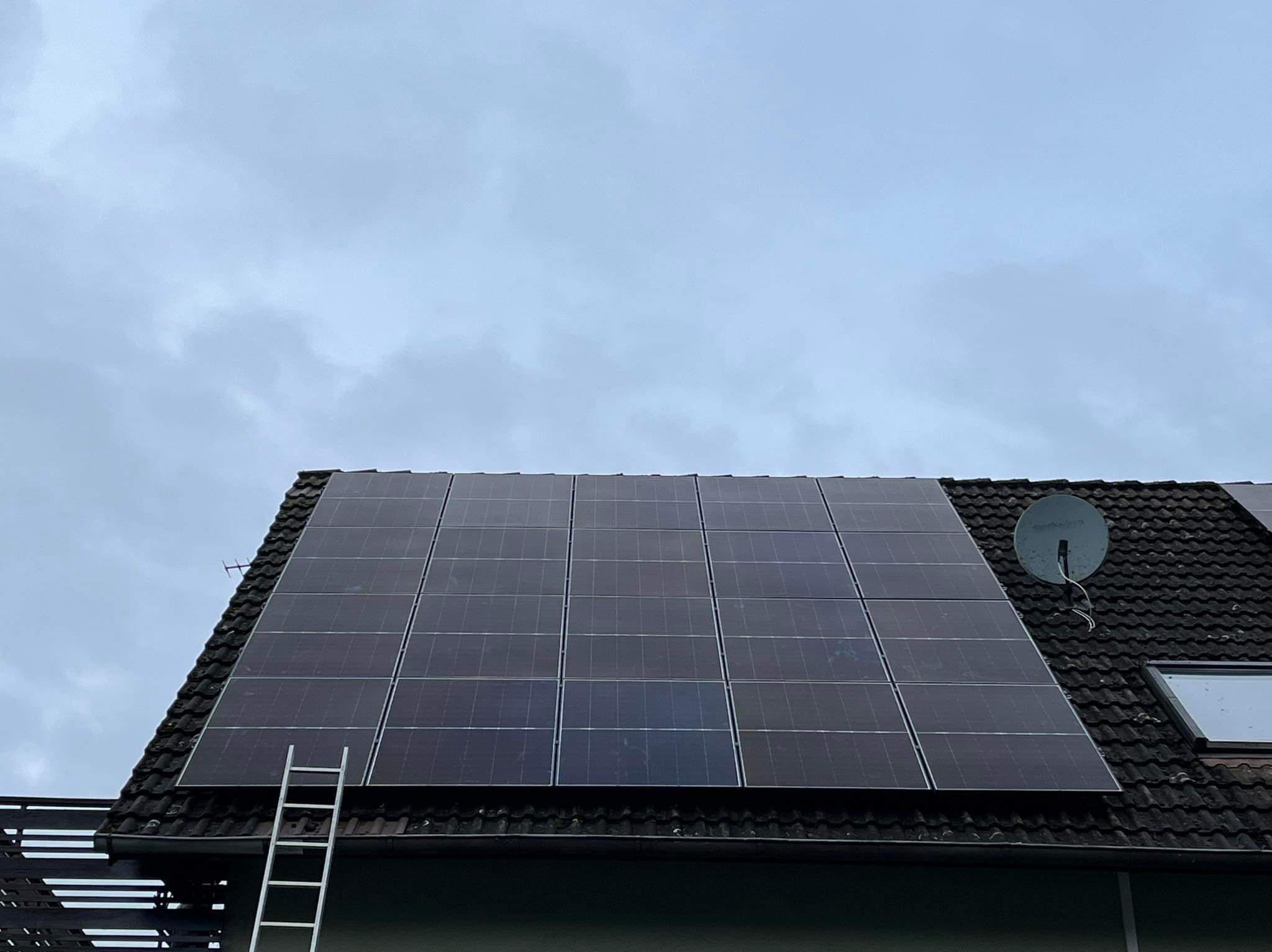 Panele słoneczne na ciemnym, spadzistym dachu z anteną satelitarną, drabina oparta o rynnę, pochmurne niebo w tle.