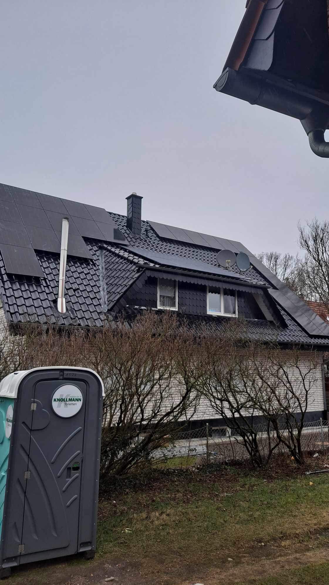 Dom z ciemną dachówką i panelami słonecznymi, widoczna instalacja wentylacyjna i dwie anteny satelitarne, na pierwszym planie przenośna toaleta z logo firmy.