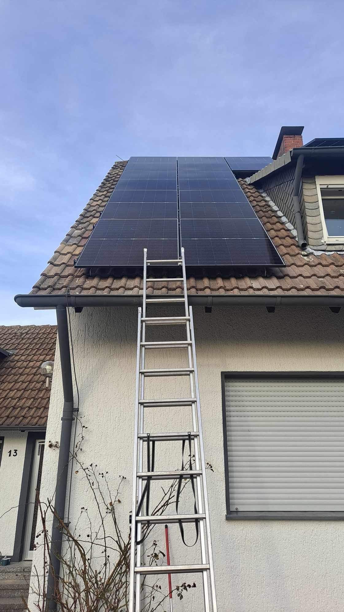 Panele słoneczne na brązowym dachu domu jednorodzinnego, drabina oparta o ścianę, widoczne okno z roletą i numer domu 13.