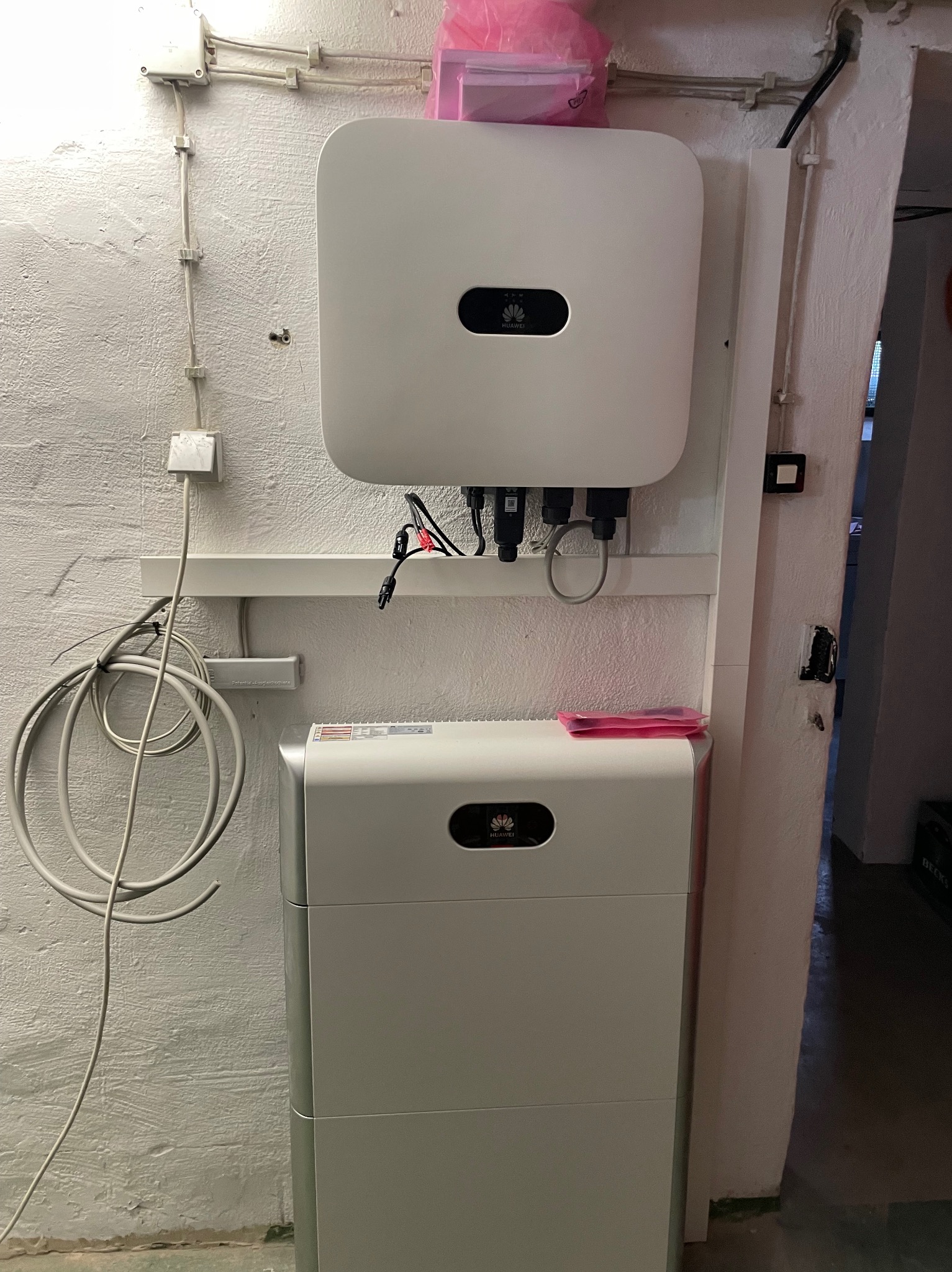 Biały falownik Huawei i moduł magazynowania energii zamontowane na pomalowanej na biało ścianie z widocznymi przewodami i elementami instalacji elektrycznej.